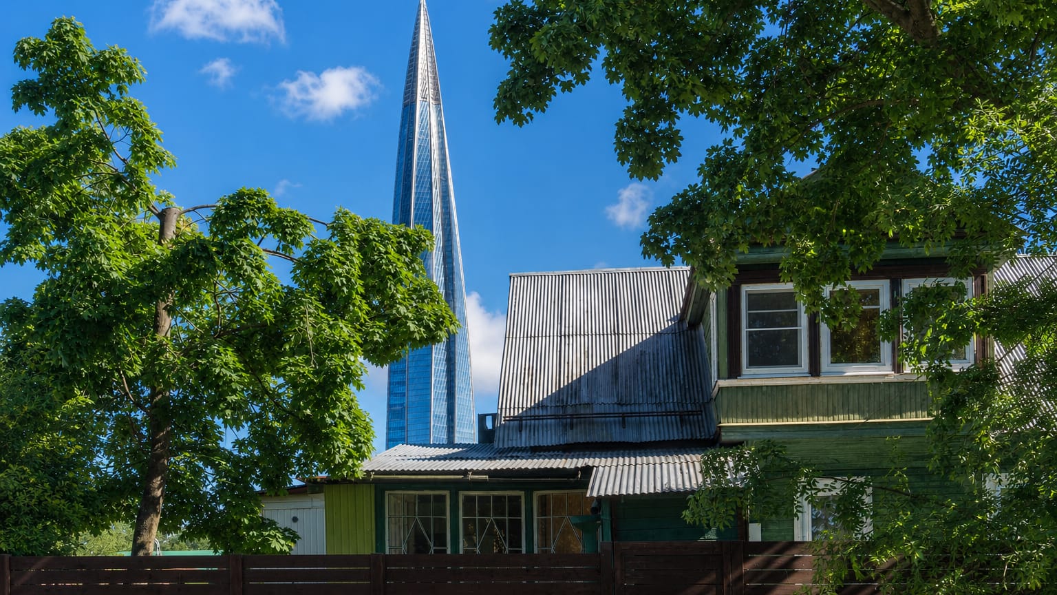 The Lakhta Center skyscraper towering behind an old wooden dacha and green trees in Lakhta, Saint Petersburg.