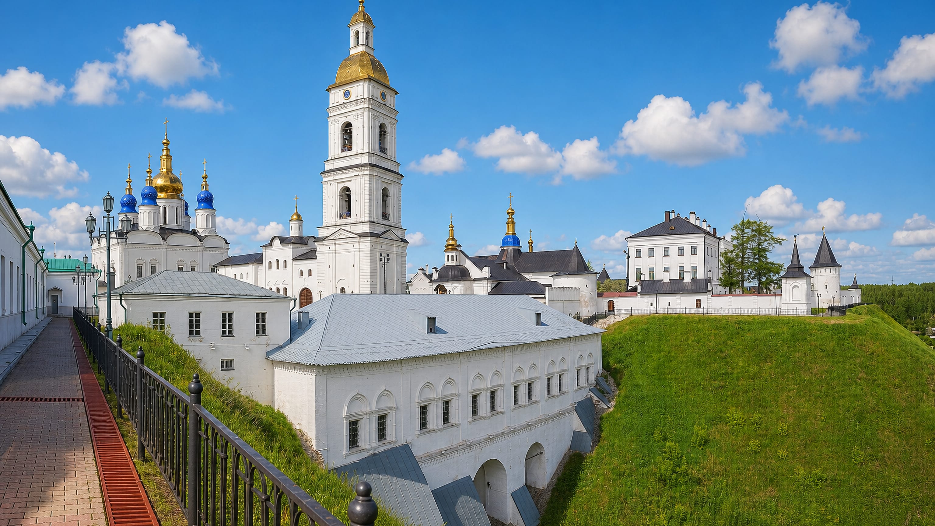 White walls, golden bell tower and blue-domed cathedral of the Tobolsk Kremlin above green ramparts, Russia.