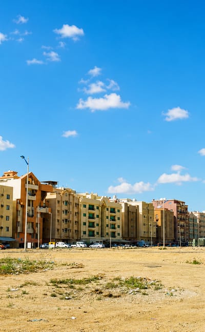 Modern glass towers rising behind beige apartment blocks across a sandy lot in Khobar