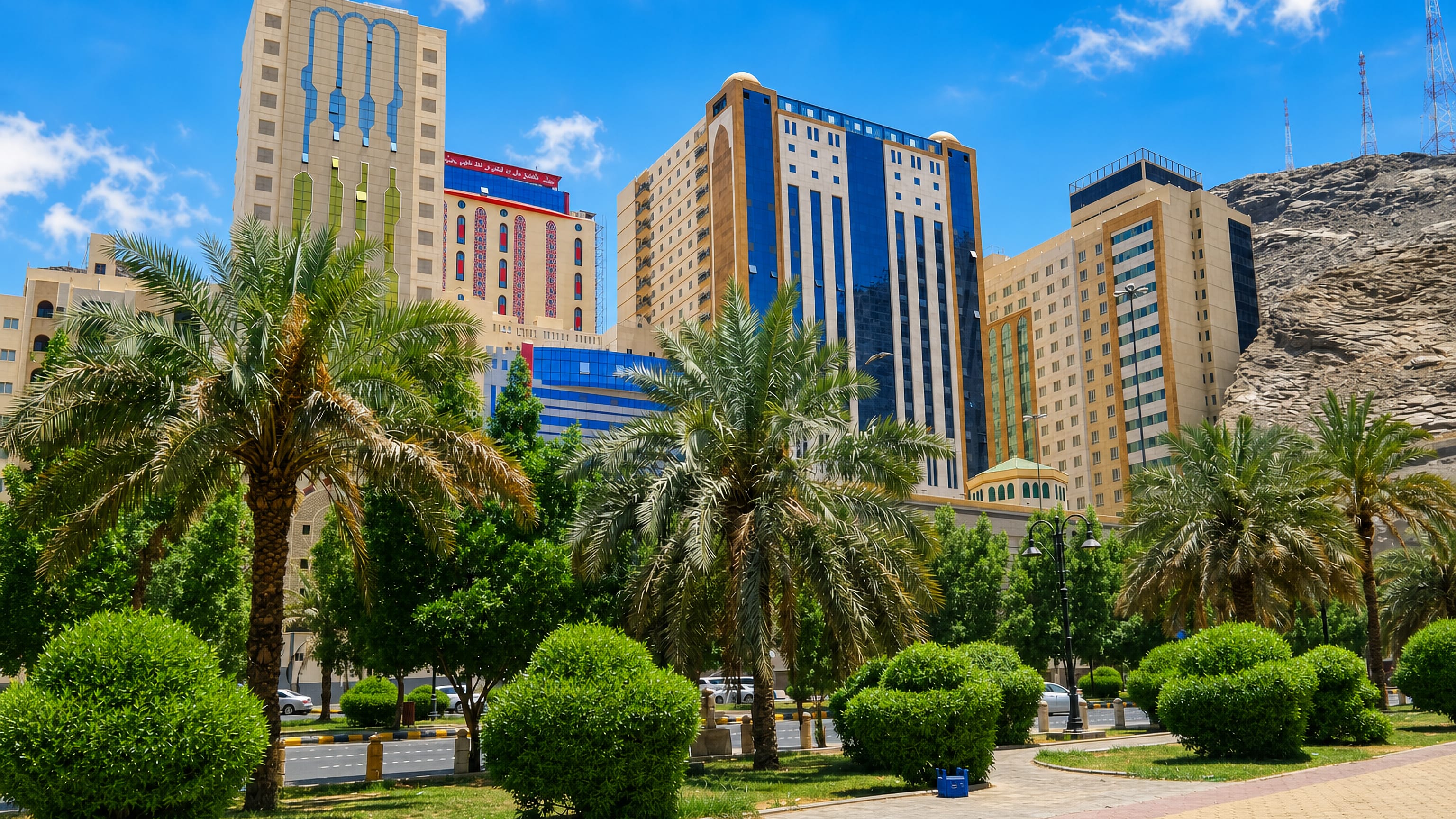 Ajyad district in Makkah with palm-lined gardens, mid-rise hotel towers and a bare rocky mountain behind under a bright sky