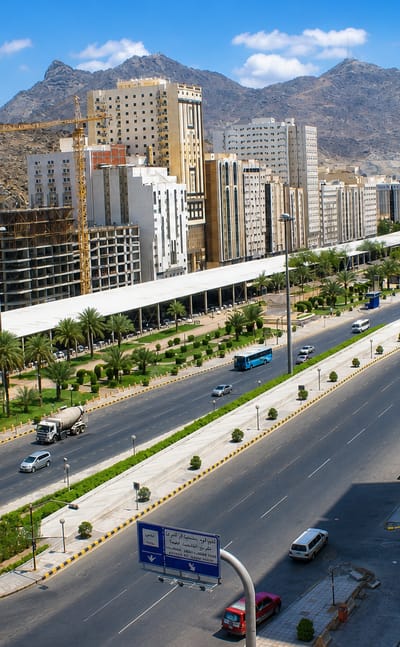 Wide daytime view of Aziziyah, Makkah, with a multi-lane highway, mid-rise buildings and dry rocky mountains in the distance