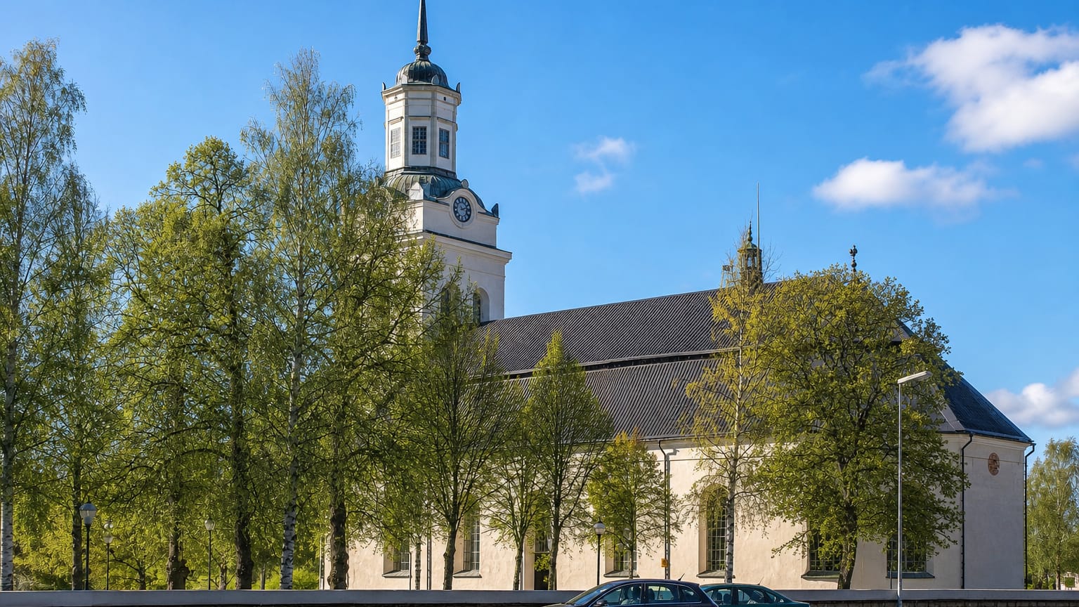 Orsa's large white church with its clock tower and cross flanked by spring birches under a blue sky.