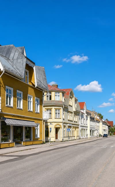 Sunny street in Vansbro lined with traditional yellow wooden houses and green trees under a blue sky.