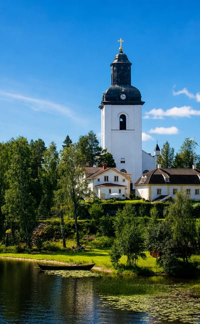 White church with a black domed tower rising above a wooded lakeshore in Ljusdal, Sweden.