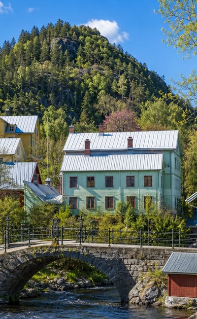 Timber houses with metal roofs and a stone arch bridge over a stream below a forested hill in Sandviken, Sweden.