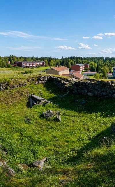 Grassy stone ruins overlooking Järpen village, its red buildings and a lake in Jämtland's forested hills.