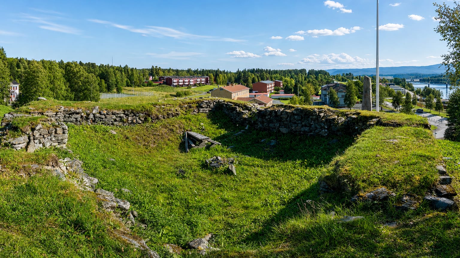 Grassy stone ruins overlooking Järpen village, its red buildings and a lake in Jämtland's forested hills.