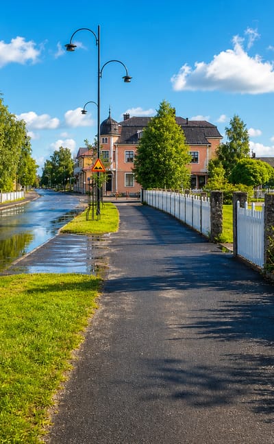 A tree-lined riverside path and street leading toward Sveg's manor-style buildings under a blue summer sky.