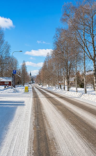 Snow-covered main street in Vemdalen lined with birches and red timber houses, a church spire in the distance.
