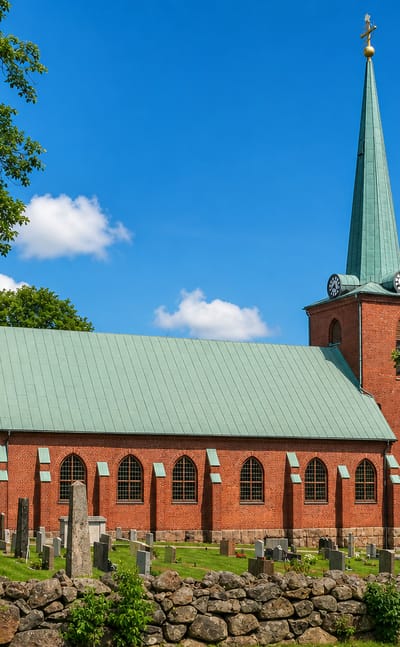 Red-brick church with a green copper spire and adjoining cemetery behind a stone wall in Gislaved, Sweden.