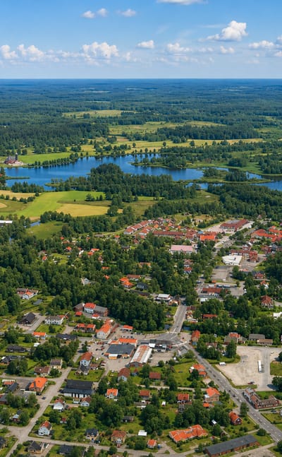 Aerial view of Savsjo, Sweden, nestled among forests and fields with a lake beyond under a summer sky.