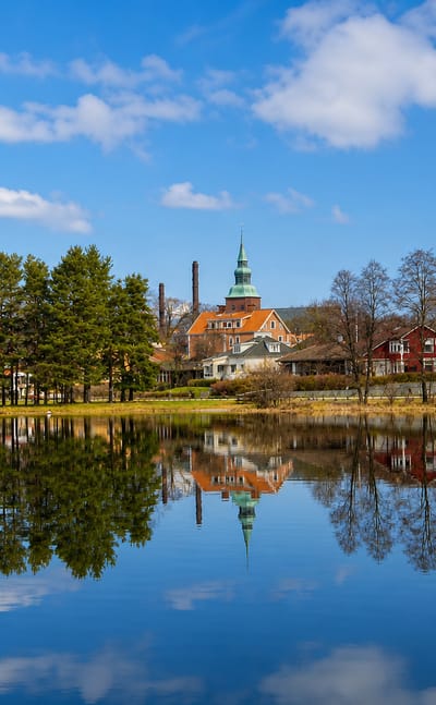 Nybro, Sweden, with a green-spired church and waterside houses mirrored in calm water under a blue sky.