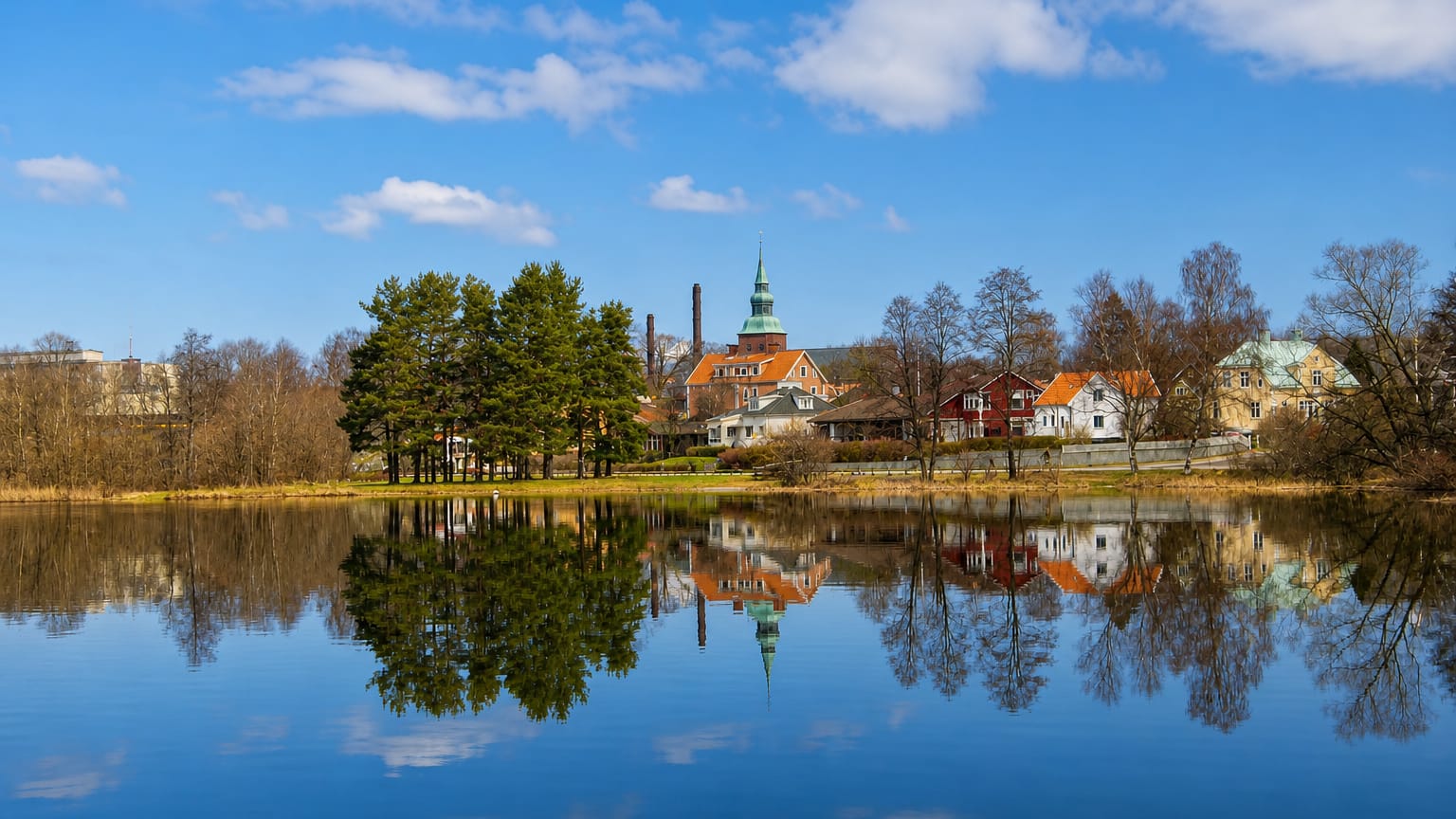 Nybro, Sweden, with a green-spired church and waterside houses mirrored in calm water under a blue sky.
