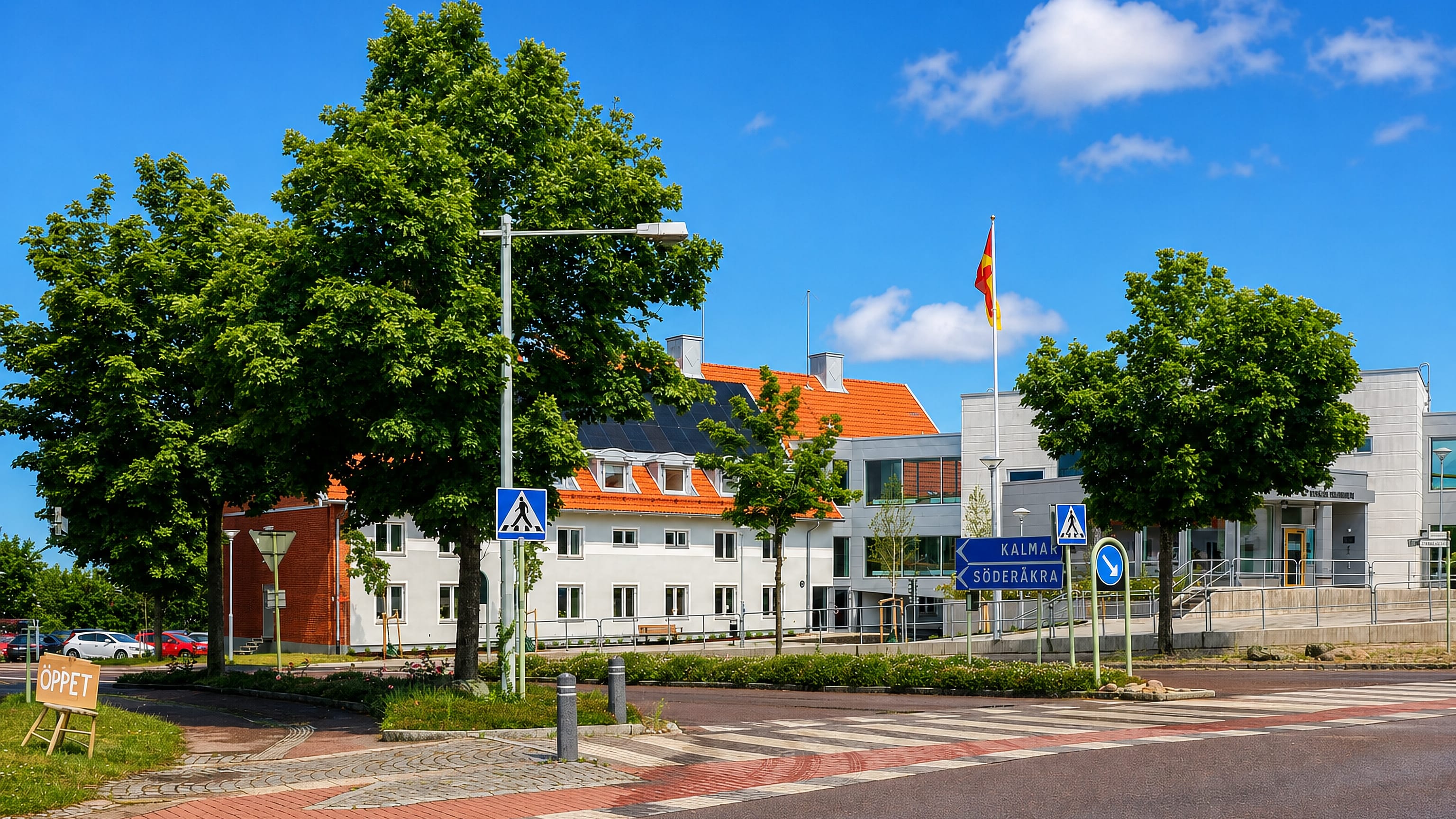 Sunny street in Torsås, Sweden, with white tile-roofed buildings, a flag, and road signs to Kalmar and Söderåkra.