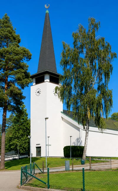 Lessebo's white modernist church with a tall dark spire and clock tower beside pine and birch trees under a blue sky.