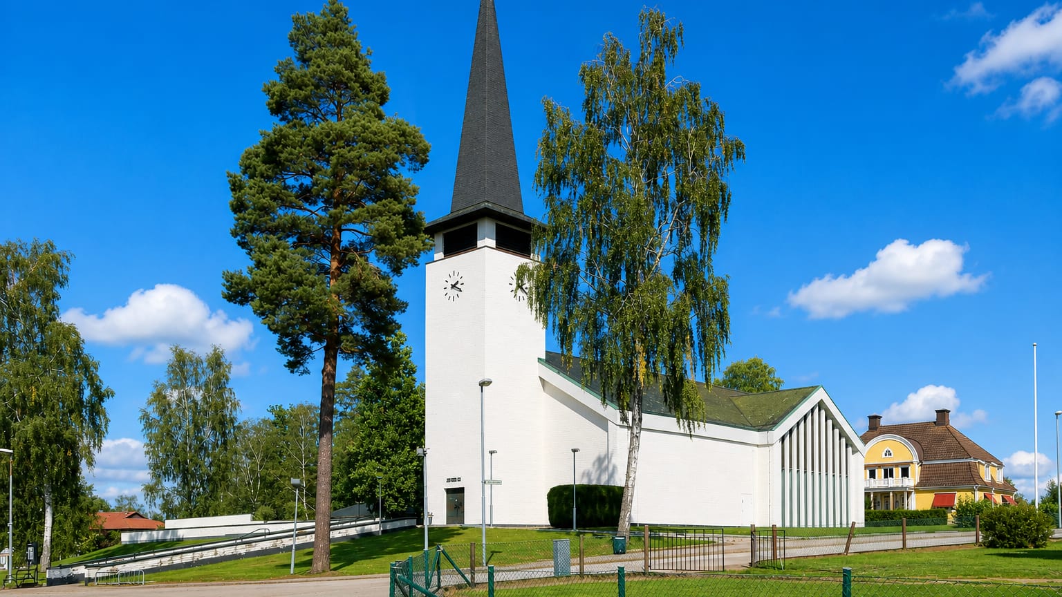 Lessebo's white modernist church with a tall dark spire and clock tower beside pine and birch trees under a blue sky.