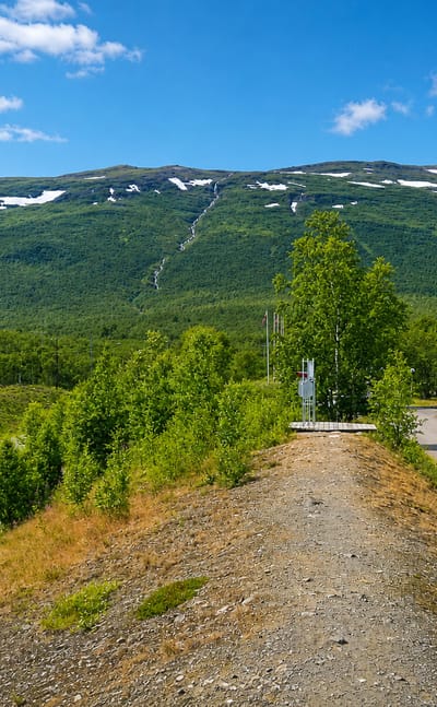 A mountain landscape at Abisko in Swedish Lapland with a road, birch scrub and a modern building below snow-streaked fells.