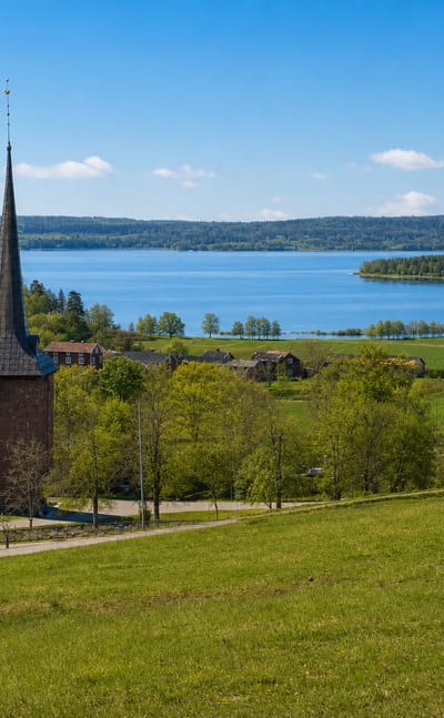 A church with a tall dark spire and red farm buildings on green meadows above a wide lake near Karlskoga, Sweden.