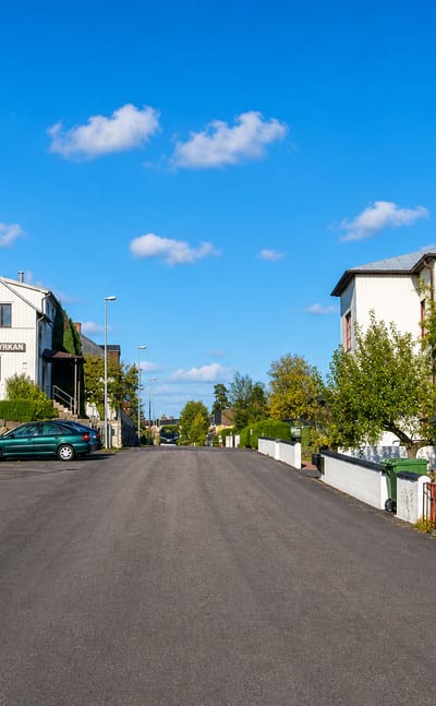 A quiet asphalt street past white timber houses and the Saron chapel in Boxholm, Sweden, under a blue sky.