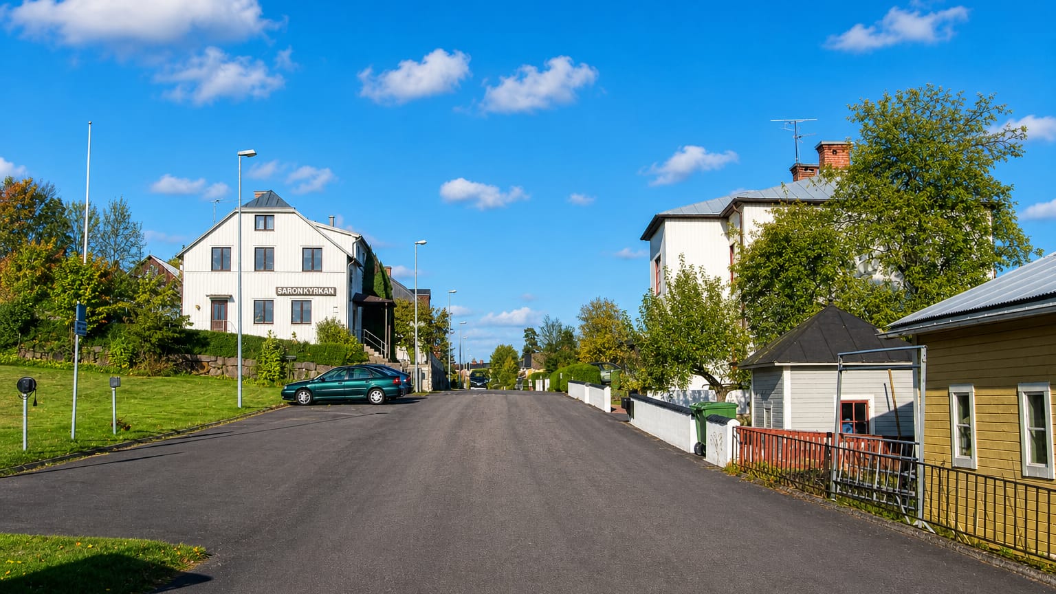 A quiet asphalt street past white timber houses and the Saron chapel in Boxholm, Sweden, under a blue sky.