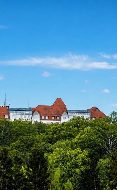 A large white building with red-tiled roofs rising above a dense green forest under a blue sky in Höör, Sweden.