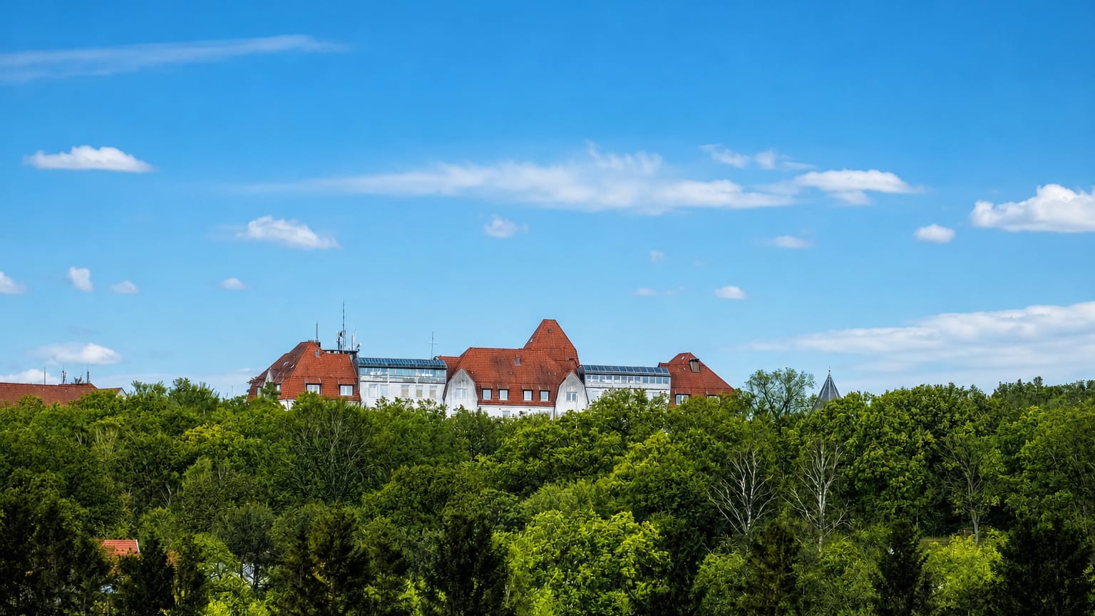 A large white building with red-tiled roofs rising above a dense green forest under a blue sky in Höör, Sweden.
