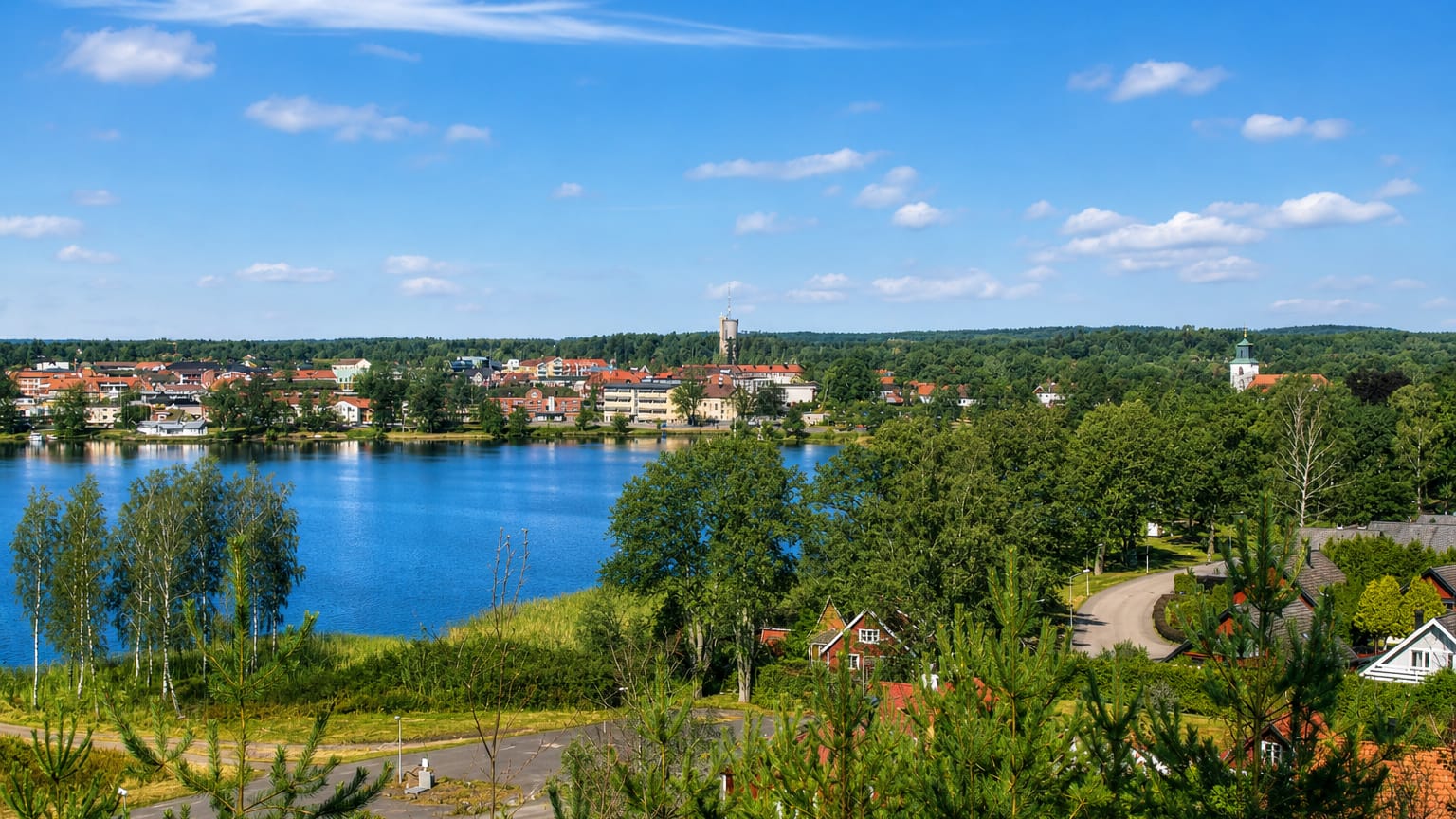 Lakeside view of Osby, Sweden, showing its water tower and church across the blue lake framed by birch trees.