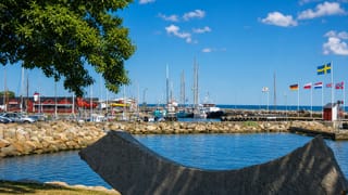 Simrishamn harbour, Sweden, with moored sailing boats, flags and a stone sculpture beside the calm blue water