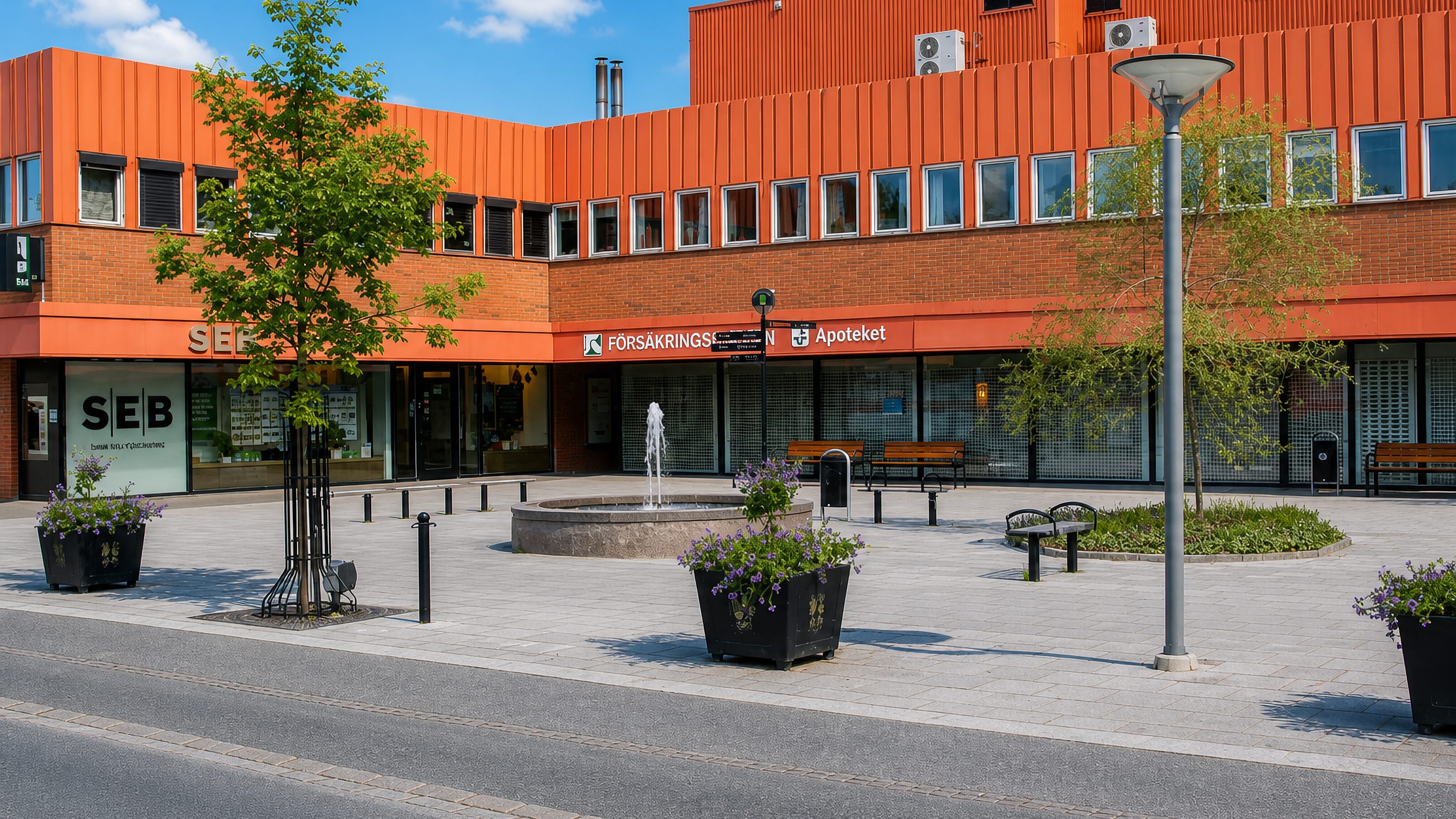 Vingåker town square with a fountain, flower planters and shops including a bank and pharmacy on a sunny day.