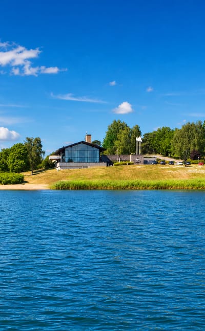 Modern glass building and a small beach on a green shoreline across blue water at Åkersberga