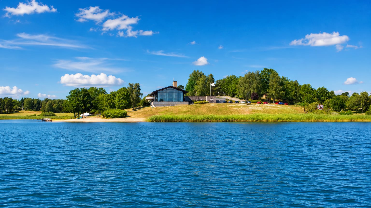 Modern glass building and a small beach on a green shoreline across blue water at Åkersberga
