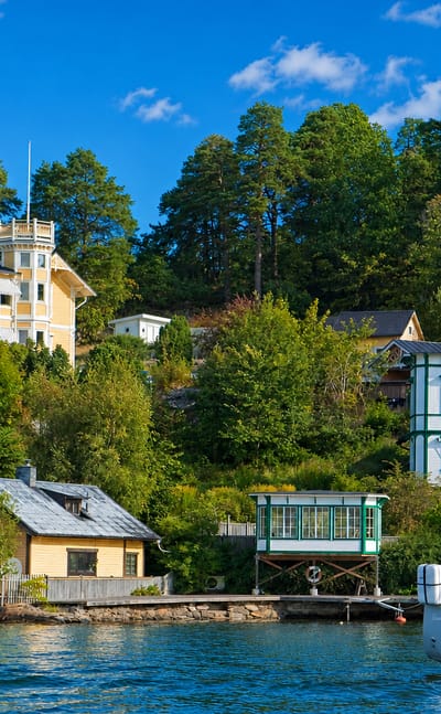 Traditional waterfront villas with a Swedish flag and a moored sailboat below a forested shore in Boo, Nacka, Sweden.