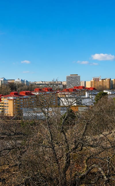 Hagalund's red-roofed apartment blocks and high-rises beyond railway tracks, viewed from a wooded hill in Solna, Sweden.