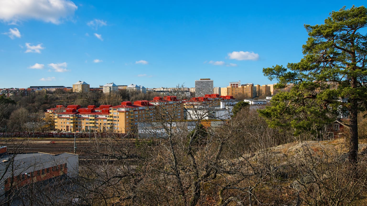 Hagalund's red-roofed apartment blocks and high-rises beyond railway tracks, viewed from a wooded hill in Solna, Sweden.