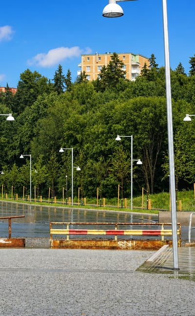 Wet paved plaza lined with street lamps in Järva, Stockholm, with modernist apartment blocks set among green wooded hills.