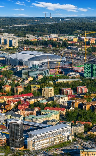 Aerial daytime view over Råsunda in Solna, Sweden, showing its large arena, colourful low-rise housing and forested lakes beyond.