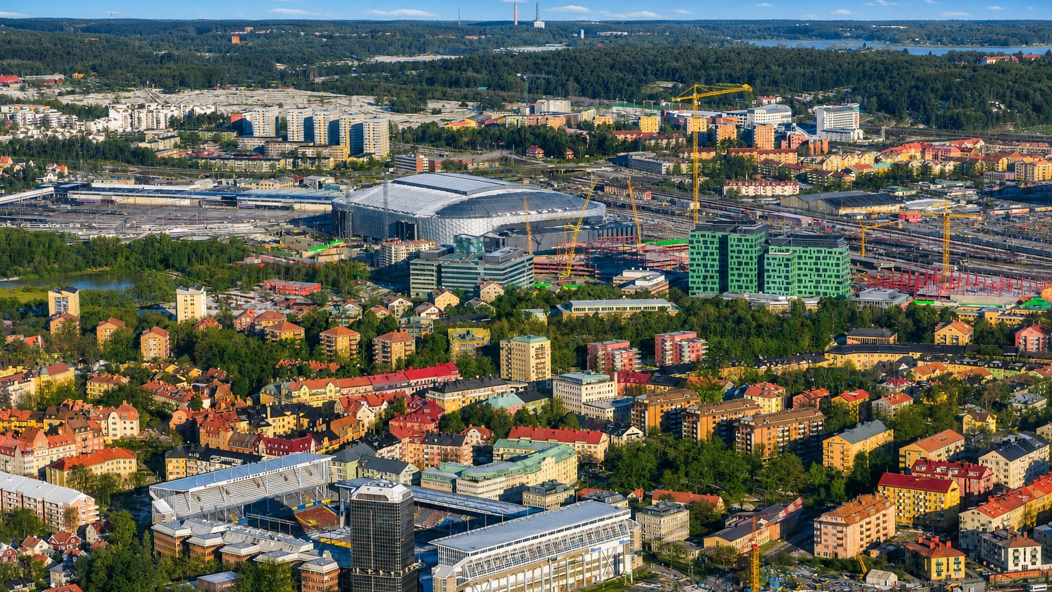Aerial daytime view over Råsunda in Solna, Sweden, showing its large arena, colourful low-rise housing and forested lakes beyond.