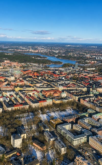 Aerial view over Vasastaden's dense grid of red-roofed apartment blocks and parks, with Stockholm's lakes beyond.