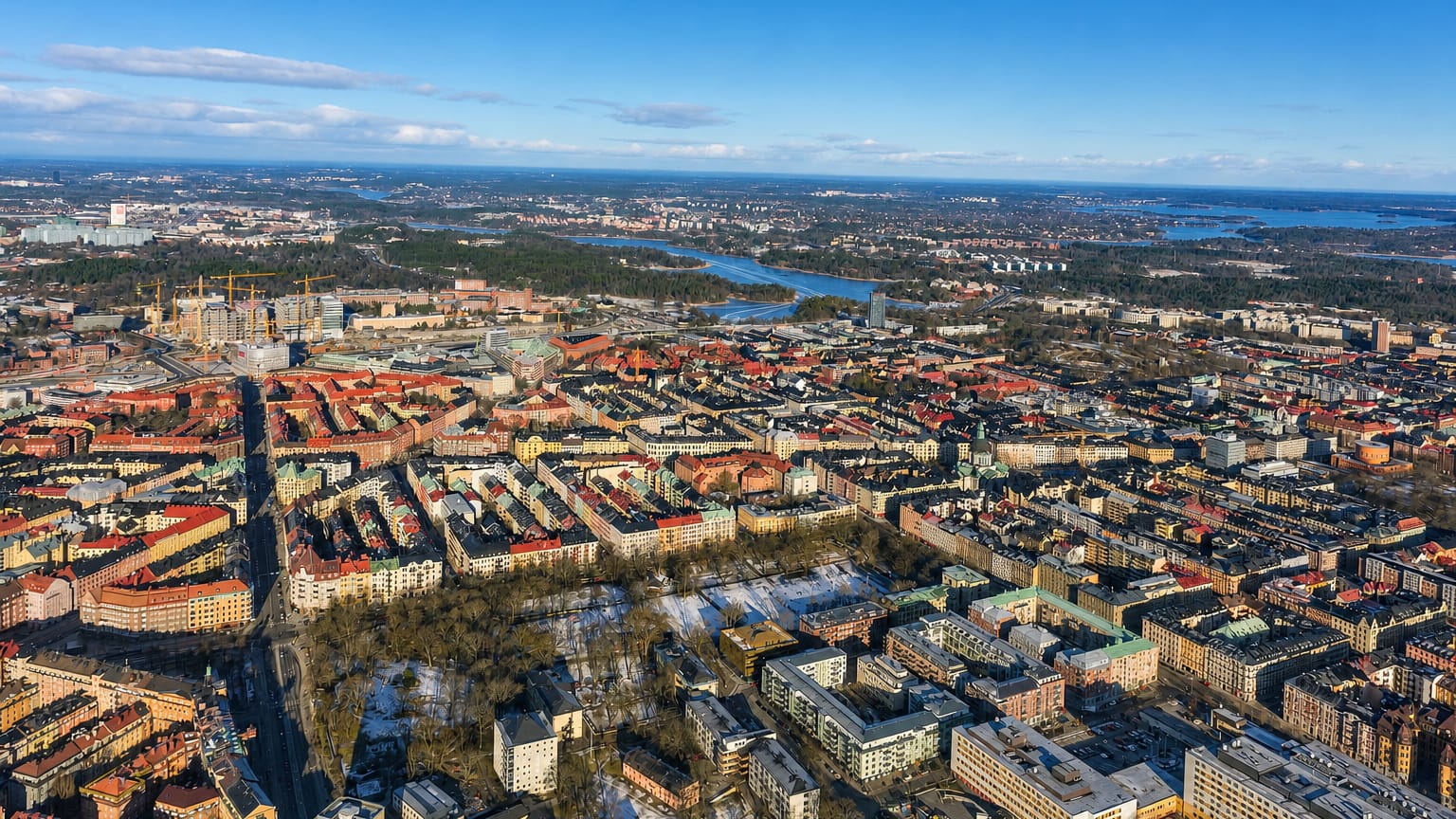 Aerial view over Vasastaden's dense grid of red-roofed apartment blocks and parks, with Stockholm's lakes beyond.