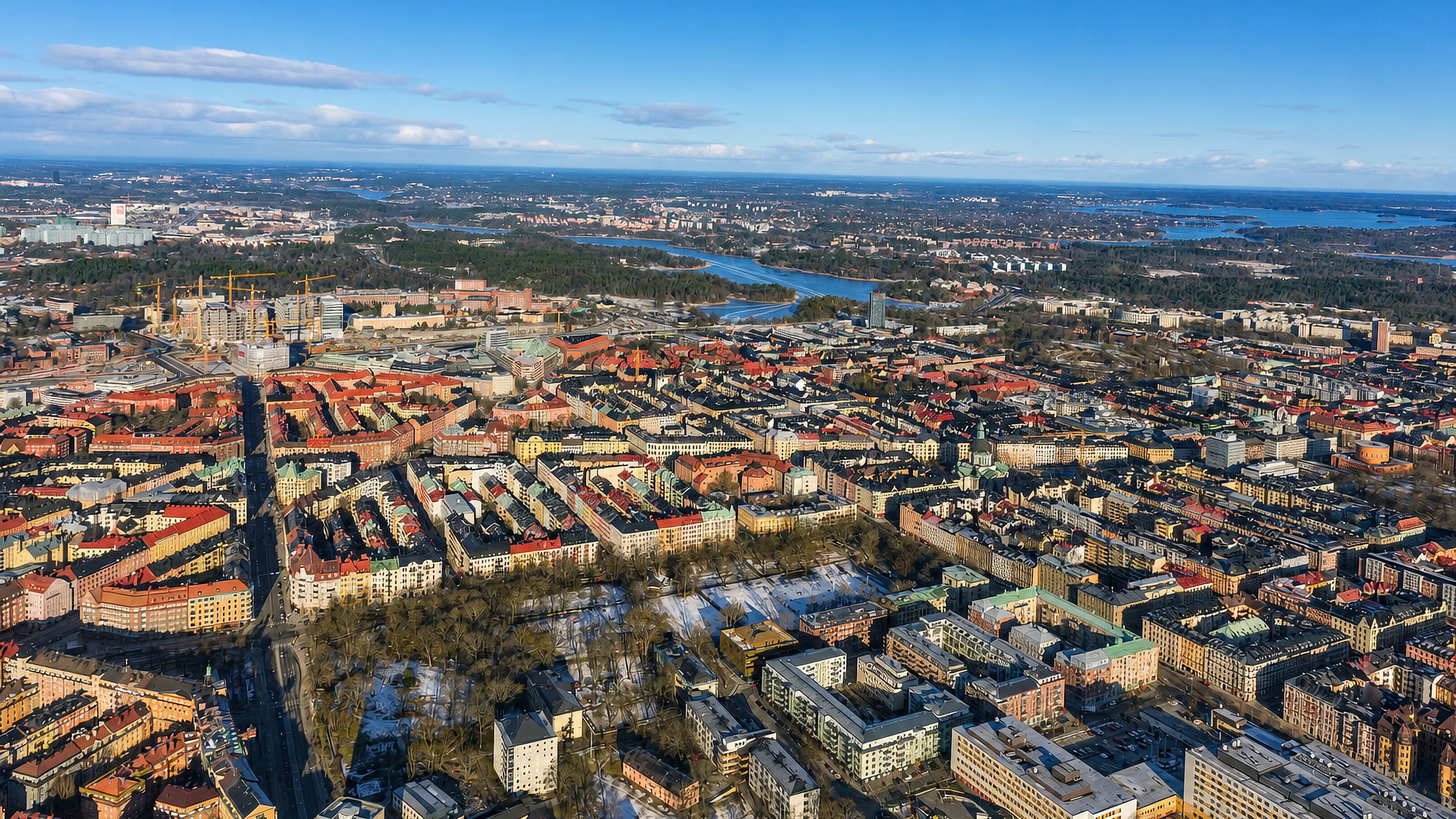 Aerial view over Vasastaden's dense grid of red-roofed apartment blocks and parks, with Stockholm's lakes beyond.