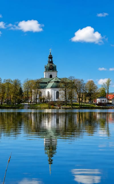 Filipstad's white church and spire reflected in a calm lake under a blue sky, with red timber houses along the wooded shore.