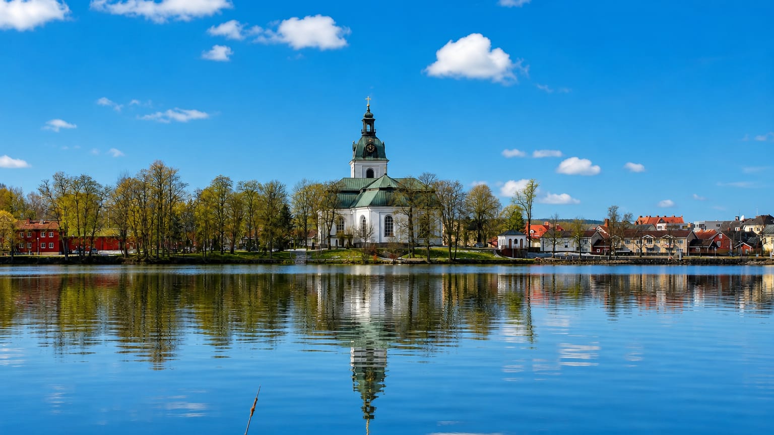 Filipstad's white church and spire reflected in a calm lake under a blue sky, with red timber houses along the wooded shore.