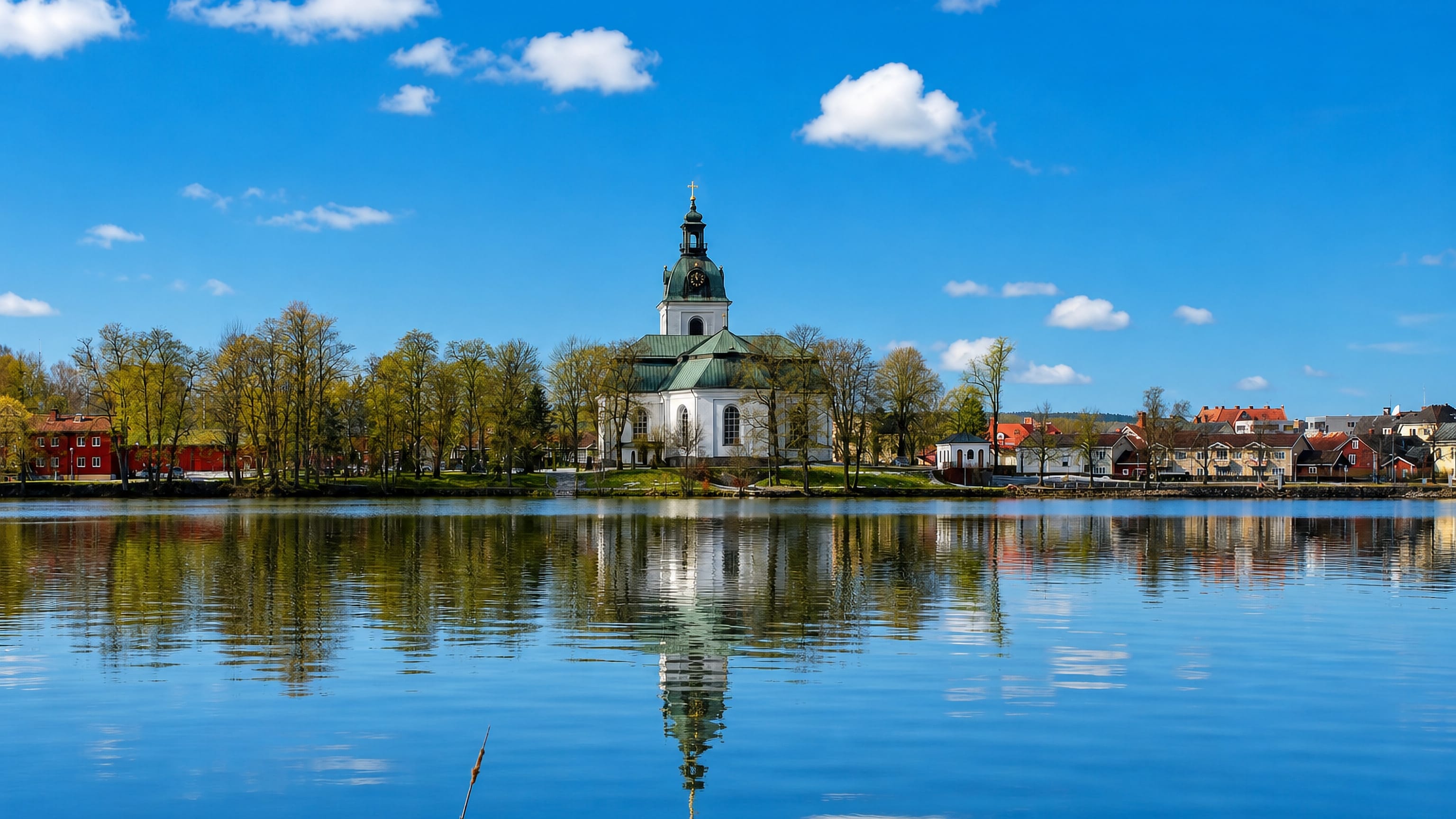Filipstad's white church and spire reflected in a calm lake under a blue sky, with red timber houses along the wooded shore.