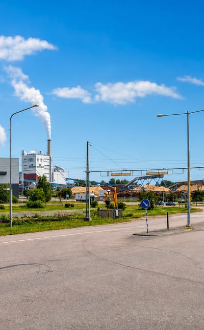 A paper mill with a steaming chimney and timber piles beside a road junction in Grums, Sweden, under a blue summer sky.