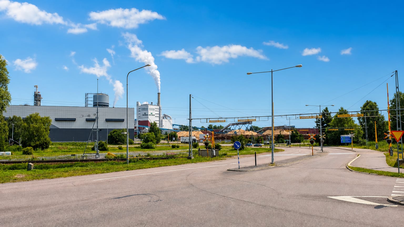 A paper mill with a steaming chimney and timber piles beside a road junction in Grums, Sweden, under a blue summer sky.