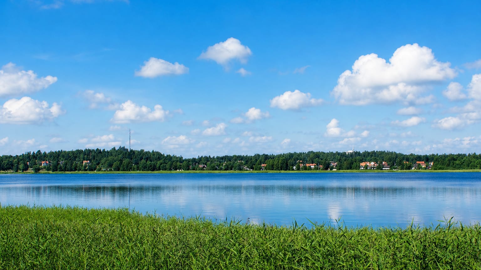 Lake Kroppkärrssjön at Kroppkärr, Sweden: reedy shore, forested far bank with scattered houses under a blue summer sky.