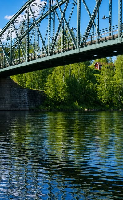 Green steel truss bridge over a river lined with birch woods and red cabins at Munkfors, Sweden.