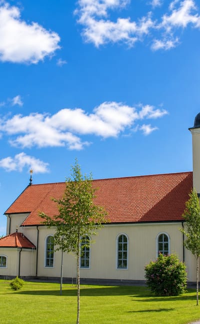 Bjurholm's pale wooden church with a red-tiled roof and black domed bell tower on a green tree-lined lawn.
