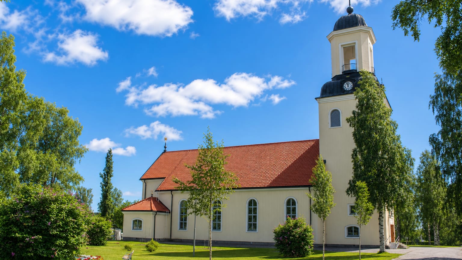 Bjurholm's pale wooden church with a red-tiled roof and black domed bell tower on a green tree-lined lawn.