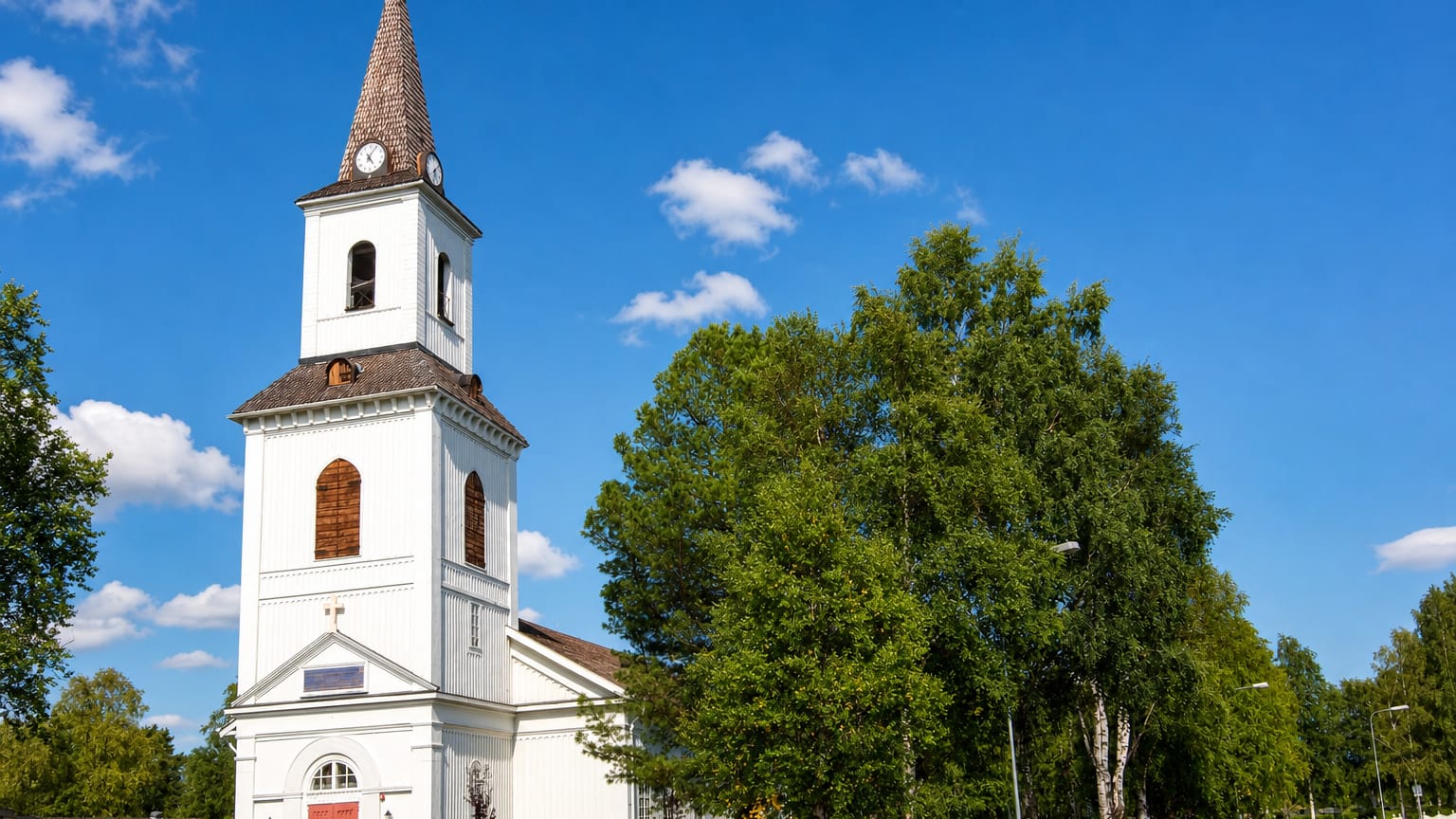 Sorsele's white wooden church with its brown shingled spire and red door beside summer birch trees under a blue sky.
